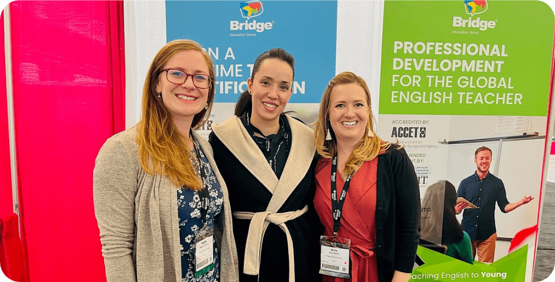 Three smiling women are standing in front of a booth display. The booth features banners with the text "Bridge Education Group" and "Professional Development for the Global English Teacher." The women are wearing conference lanyards and badges.